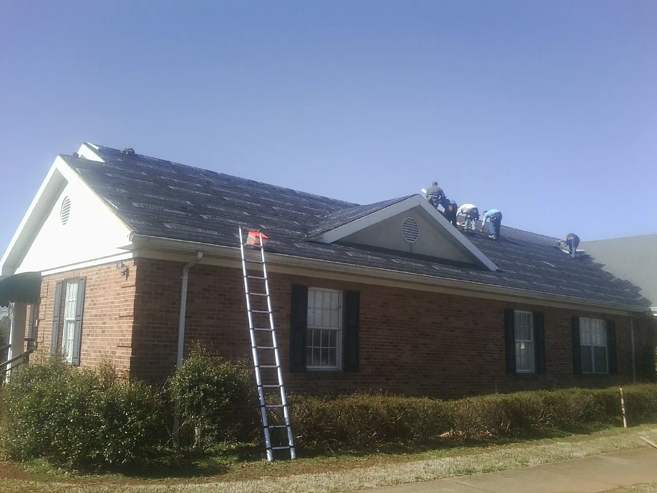 Crew tearing shingles off a brick ranch roof with a ladder set — Emergency Roof Repair work by our crew in Albia