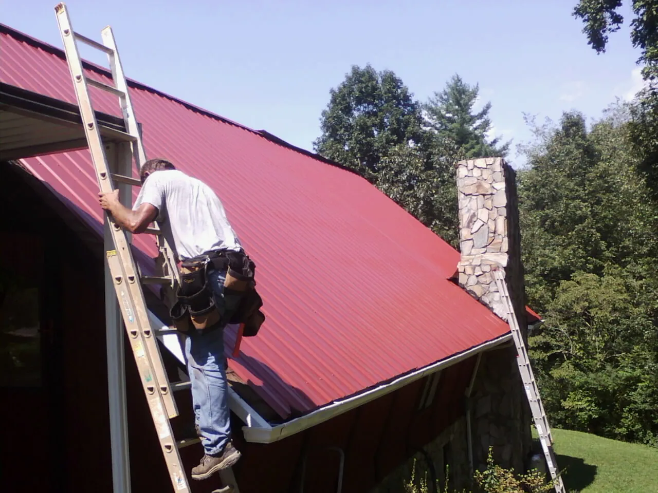 Roofer on a ladder beside a red metal roof and stone chimney — Soffit & Fascia Repair by Roofing Brothers in Albia